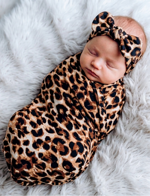 Newborn baby wrapped in a leopard print swaddle and headband on a white fluffy blanket